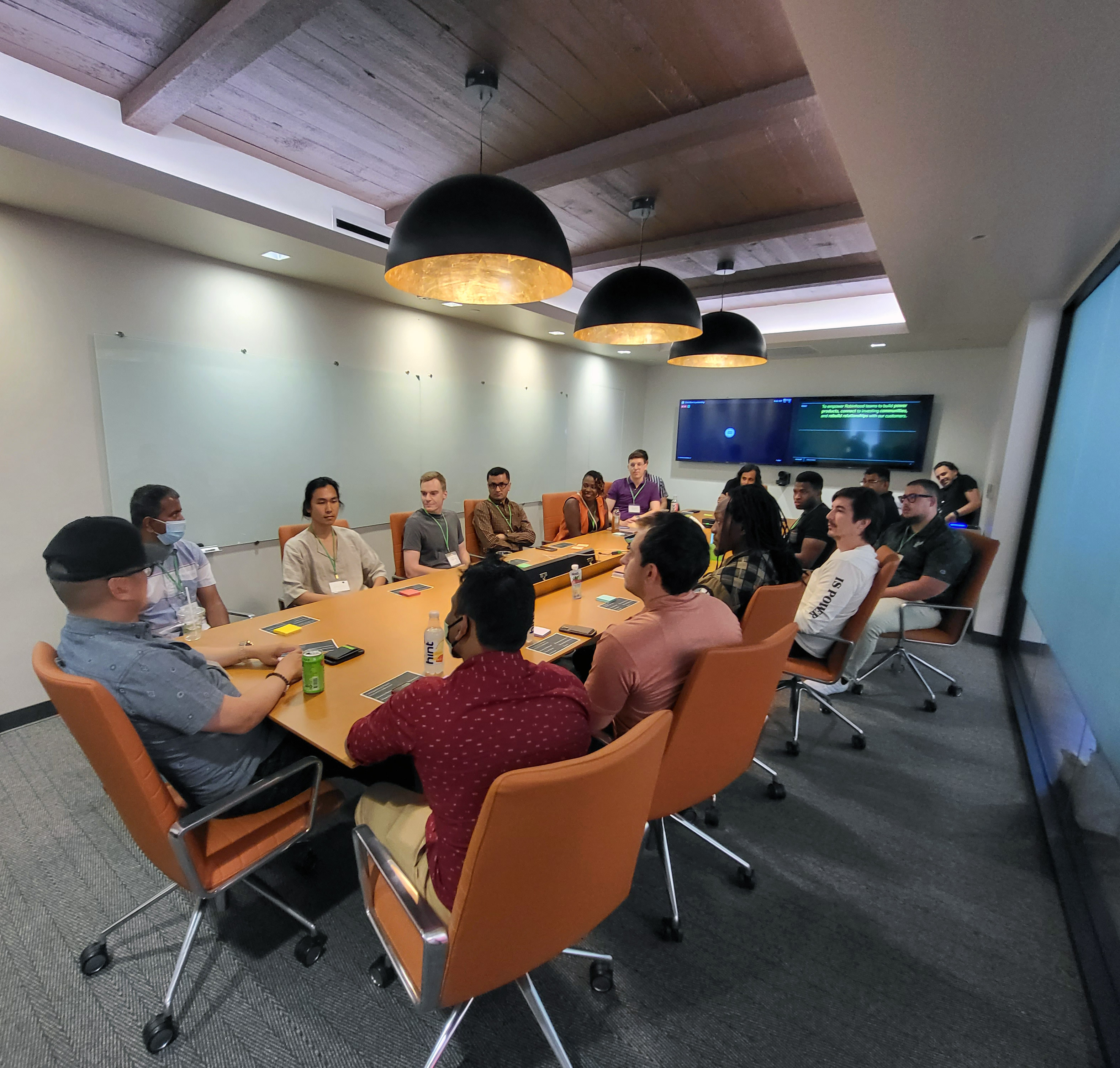 People sit in orange chairs around a long table beneath three black pendant lights in a meeting room.