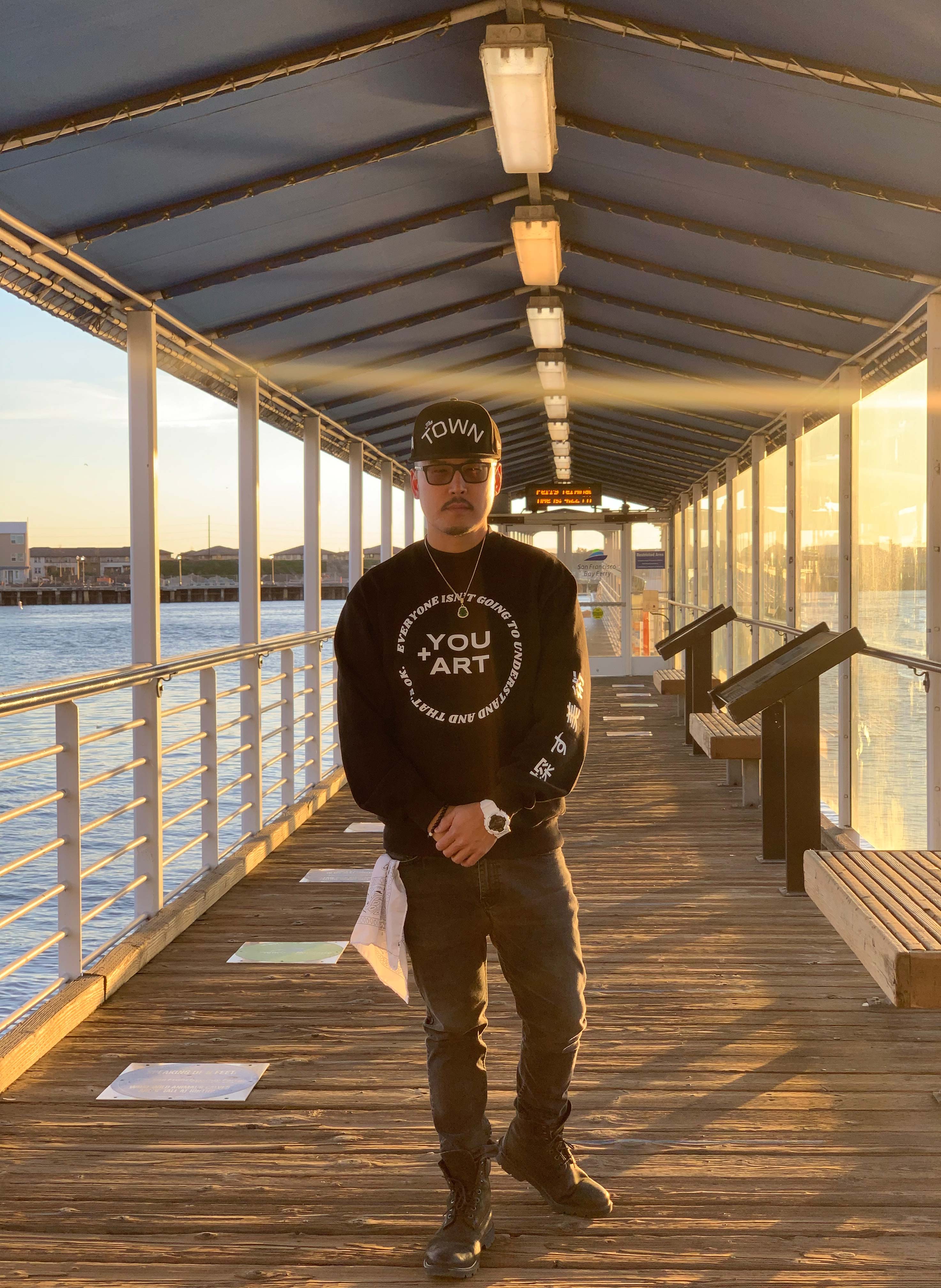 Person in a black cap and sweatshirt stands on a covered wooden pier with railings, benches and water alongside.