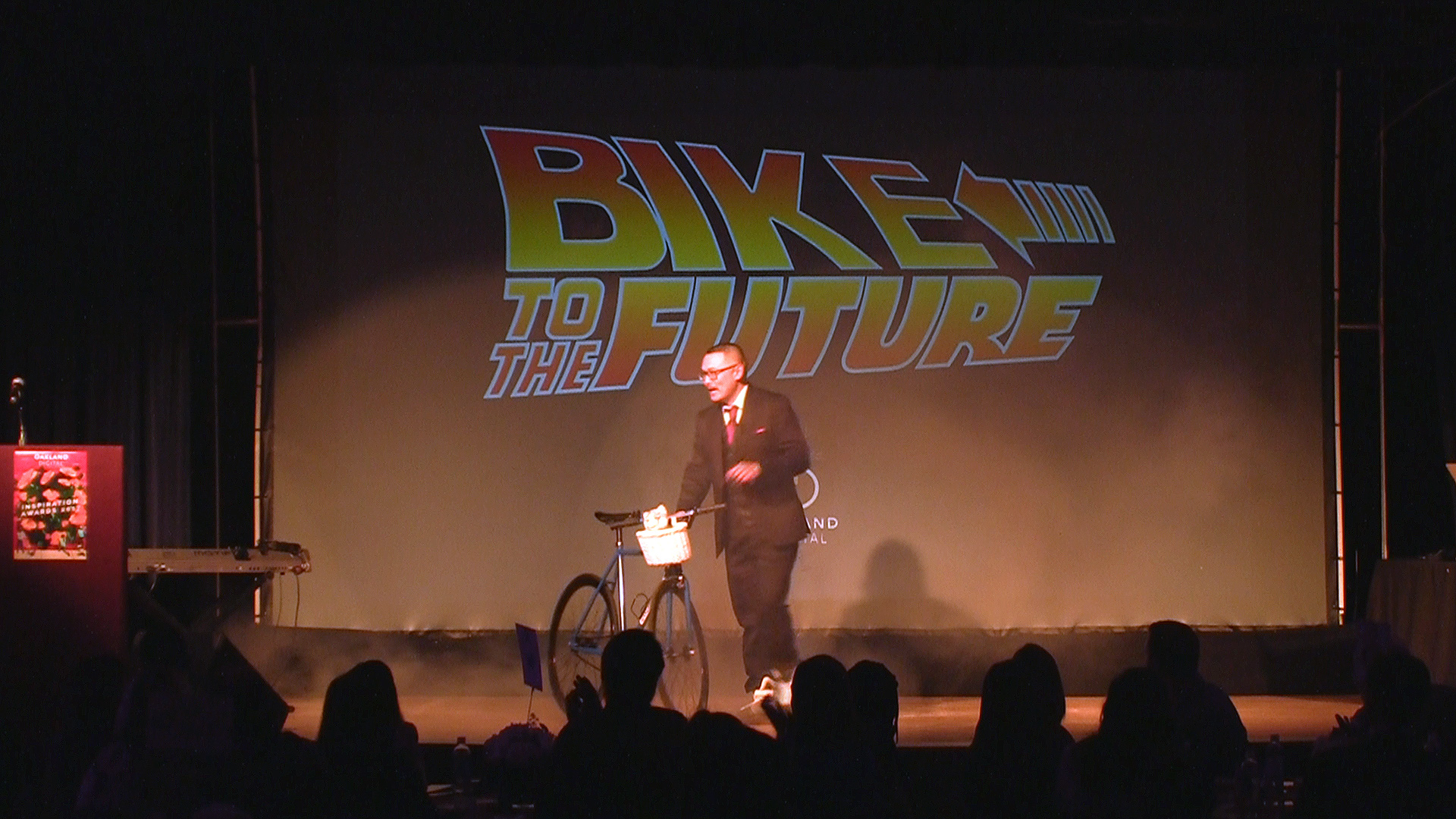 Shaun Tai stands beside a bicycle onstage beneath a “BIKE TO THE FUTURE” sign, with audience silhouettes below.