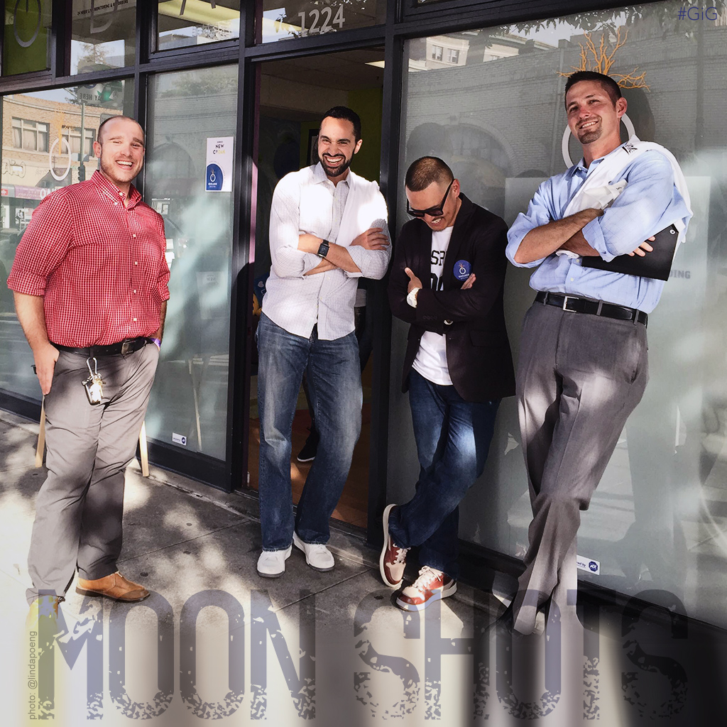 Four men pose outside a glass doorway; three have folded arms, and one looks down while the others smile.