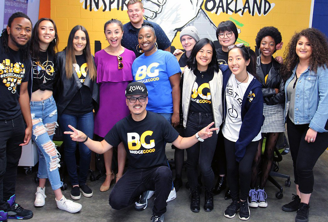 A group poses in front of a colorful painted wall, with one person kneeling in front and flashing peace signs.