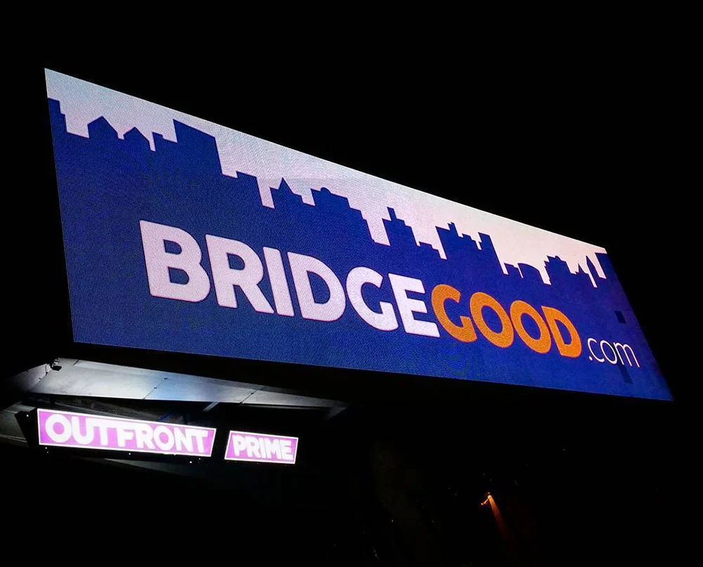 An illuminated BRIDGEGOOD.com billboard with a city skyline silhouette stands against the night sky.