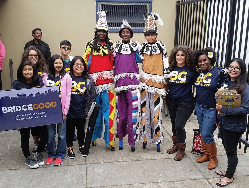 A group poses outside with three performers on stilts, several wearing BRIDGEGOOD shirts and holding a matching sign.