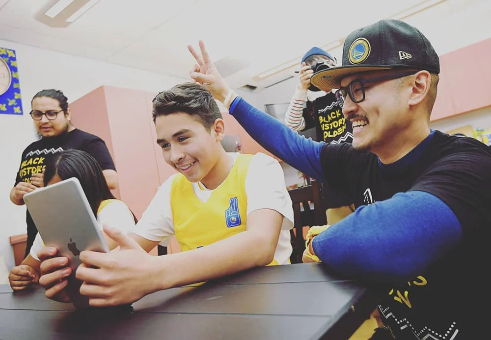 A boy holds a tablet at a classroom table as a smiling man makes bunny ears behind him, with others nearby.