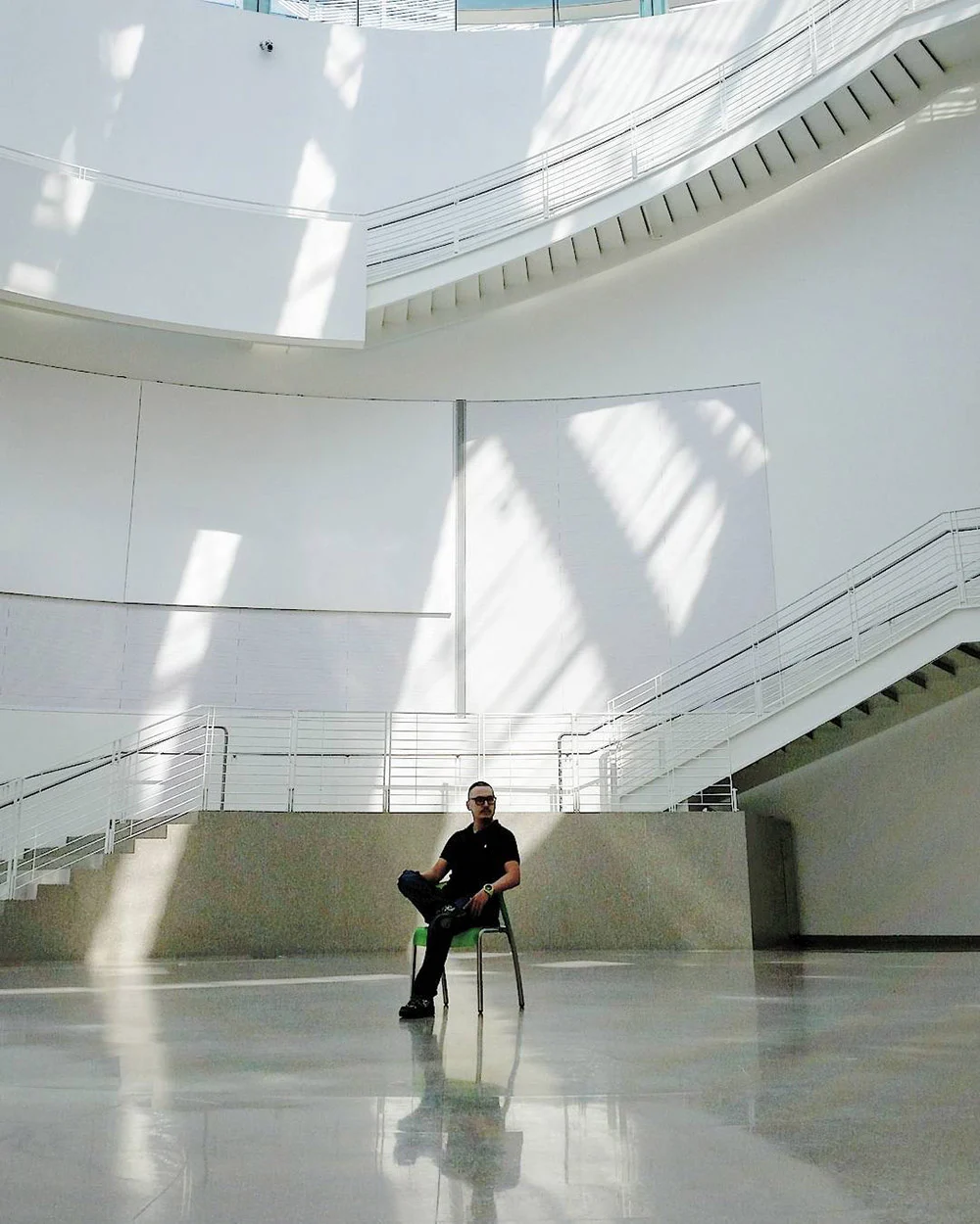 A person sits cross-legged on a chair in a bright atrium with curved ramps and sunlight across white walls.