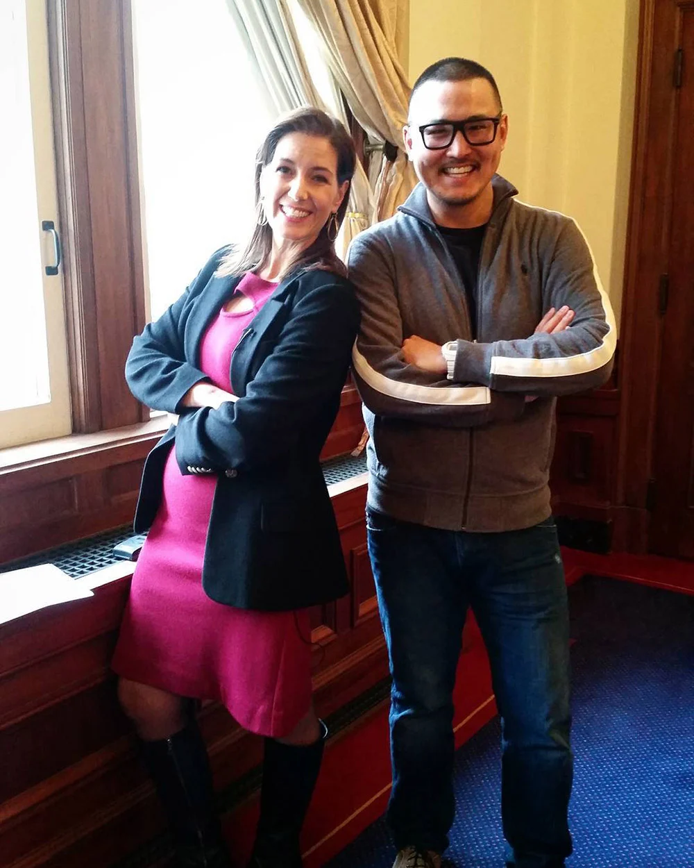 Two smiling adults stand side by side with arms crossed beside a tall window in a wood-paneled room.