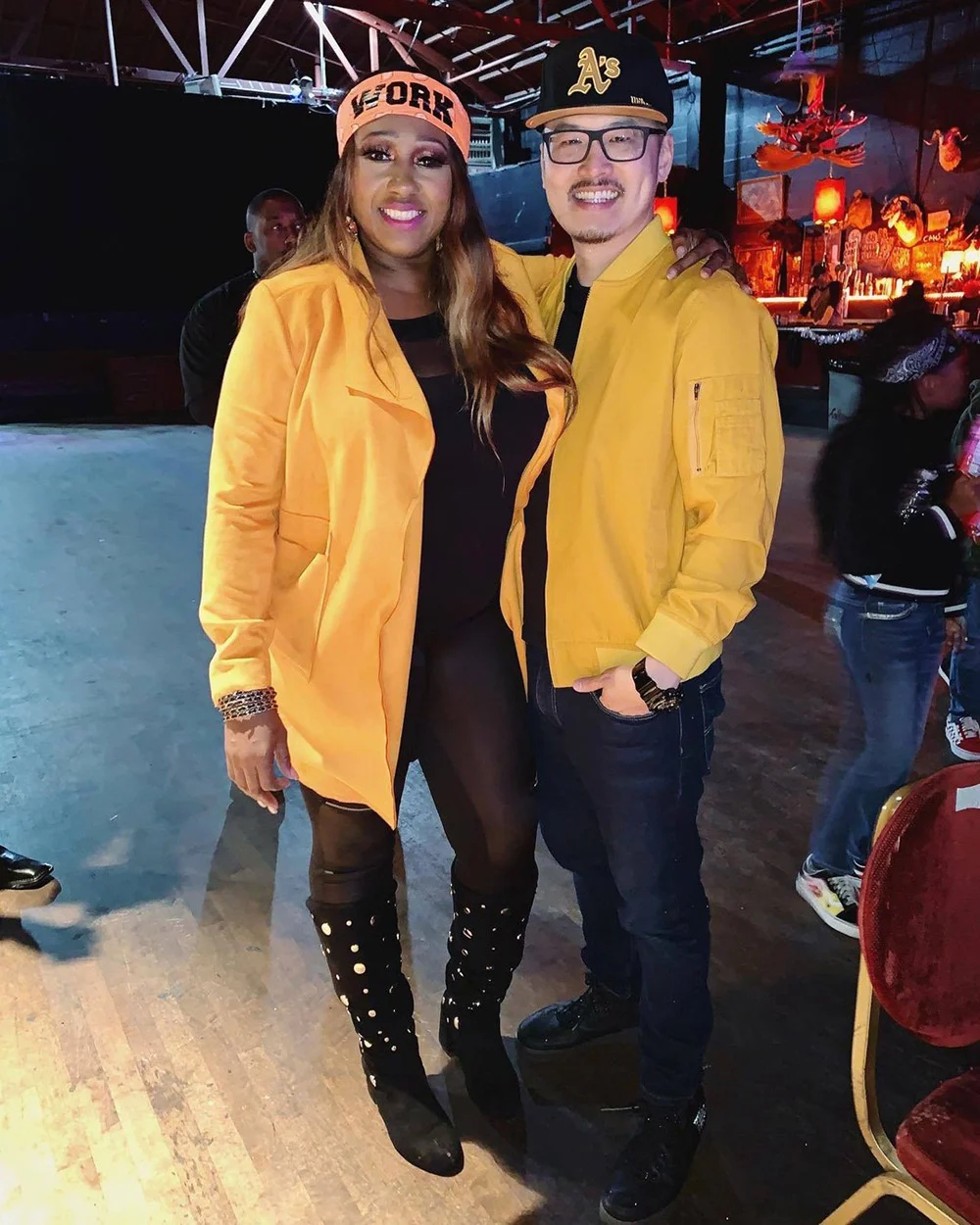 Two people in yellow jackets and baseball caps pose together in a dim venue with a glowing bar.