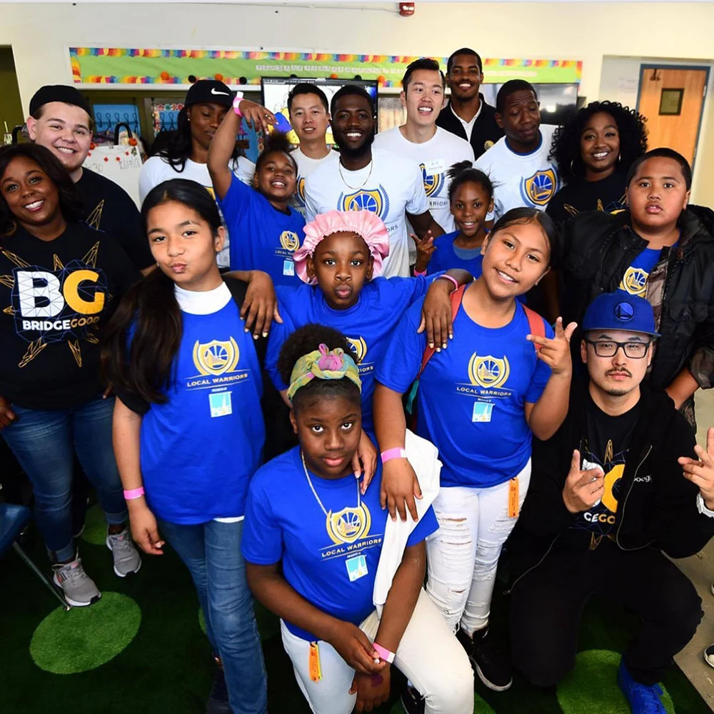 Children and adults gather for a group portrait indoors, many wearing blue and white Local Warriors shirts.