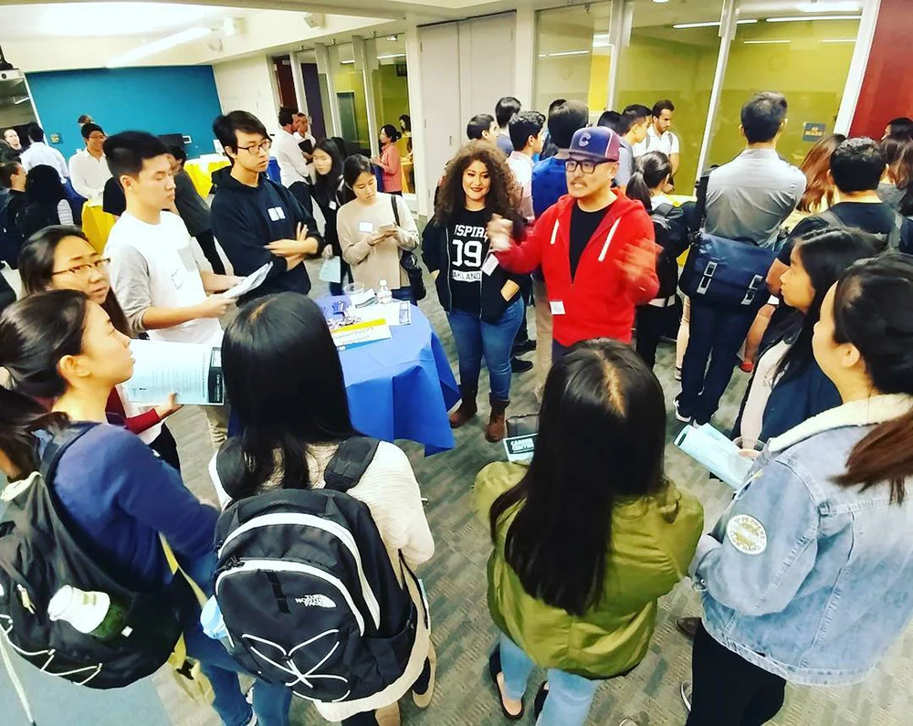 A group gathers around a person gesturing in a red jacket beside a blue-covered table in a crowded room.