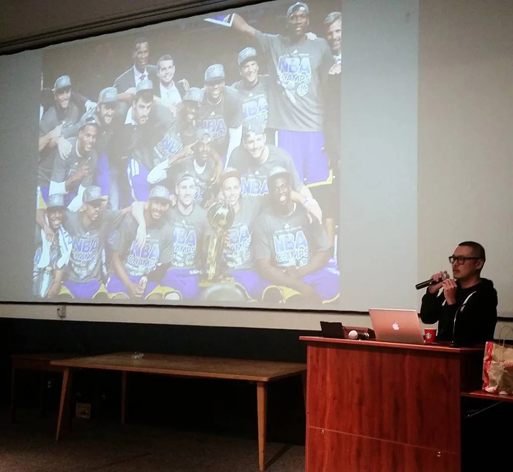 A man speaks into a microphone at a wooden podium beside a projected basketball team photo with a trophy.
