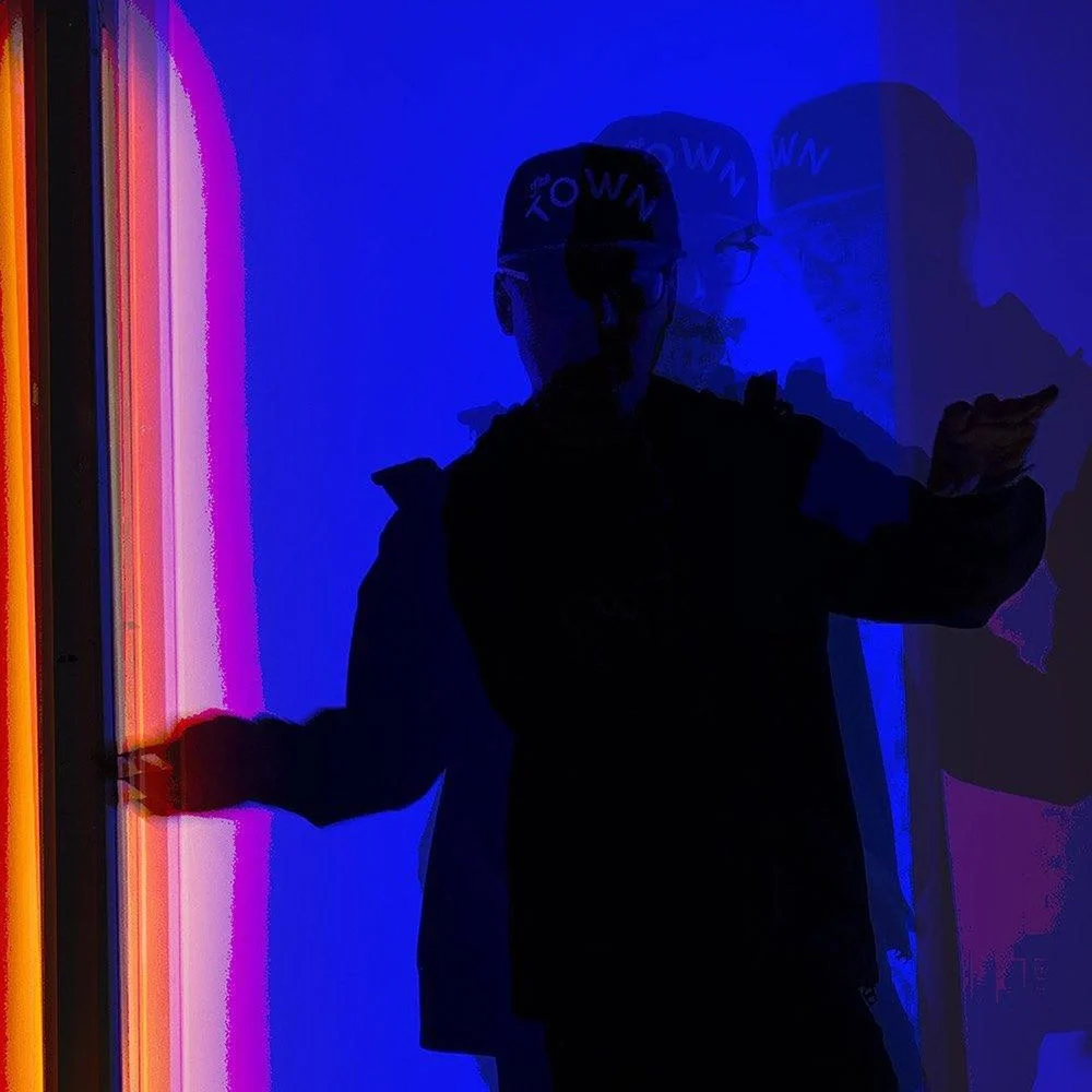 A person in a cap extends their arms beside vertical colored lights, casting overlapping shadows on a blue wall.