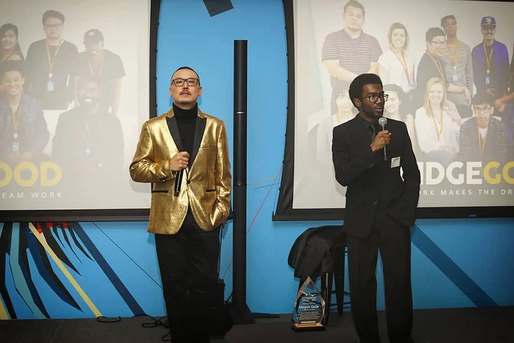 Two men hold microphones onstage, one in a gold blazer, with projected group portraits behind them.