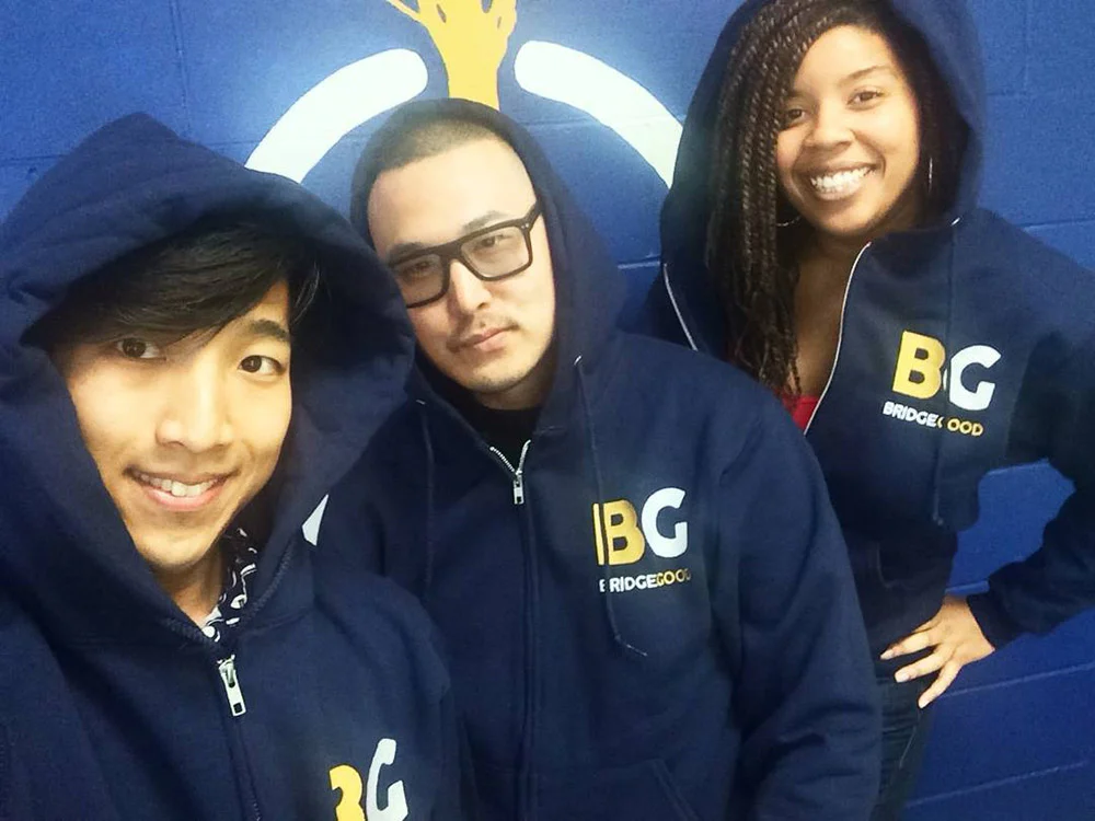 Three people pose in matching navy BRIDGEGOOD hoodies in front of a blue wall with a painted logo.