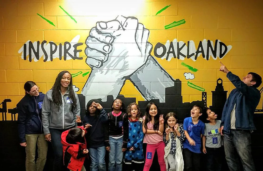 Children and adults pose before a yellow mural of clasped hands reading “Inspire Oakland,” with one person pointing up.