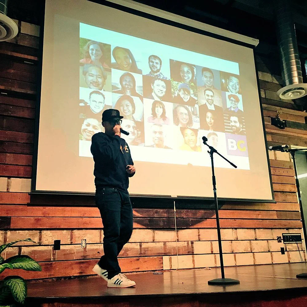 A person stands on a wooden stage beside a microphone, with a collage of faces projected behind them.