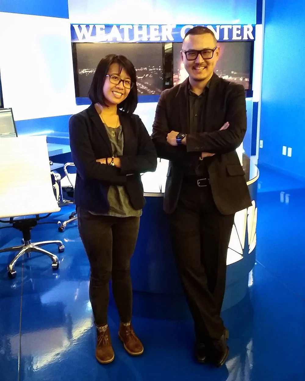 Two people wearing glasses smile with arms crossed in a blue television studio beneath a Weather Center sign.