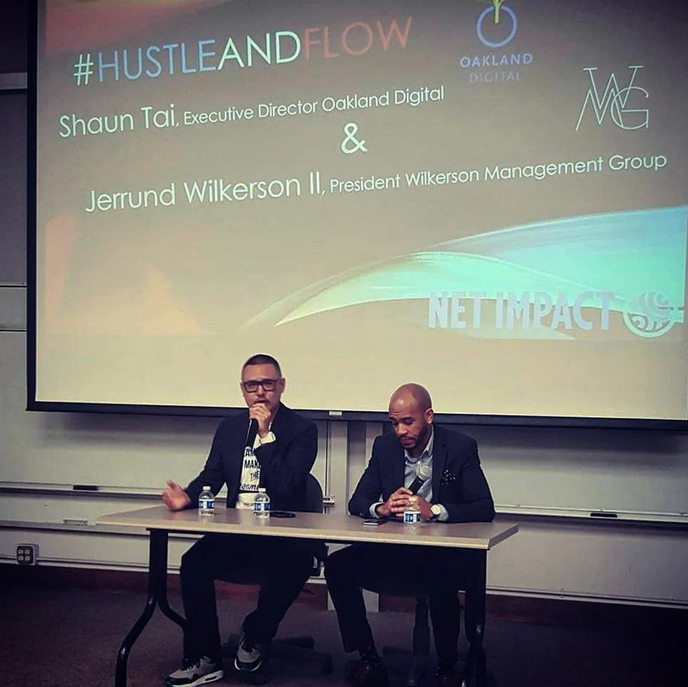 Two men sit behind a table during a panel, with a projected slide naming Shaun Tai and Jermund Wilkerson II.