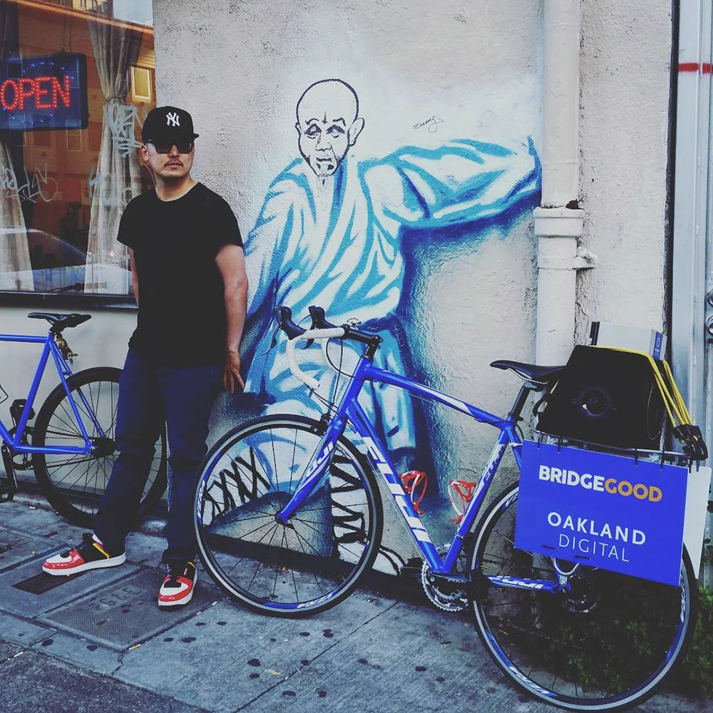 A person stands beside blue bicycles and a martial artist mural, with a BRIDGEGOOD Oakland Digital sign on one bike.