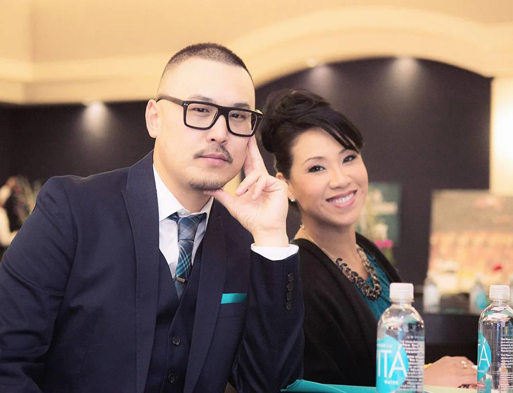 A man in glasses and a suit sits beside a smiling woman at an indoor event table with water bottles.