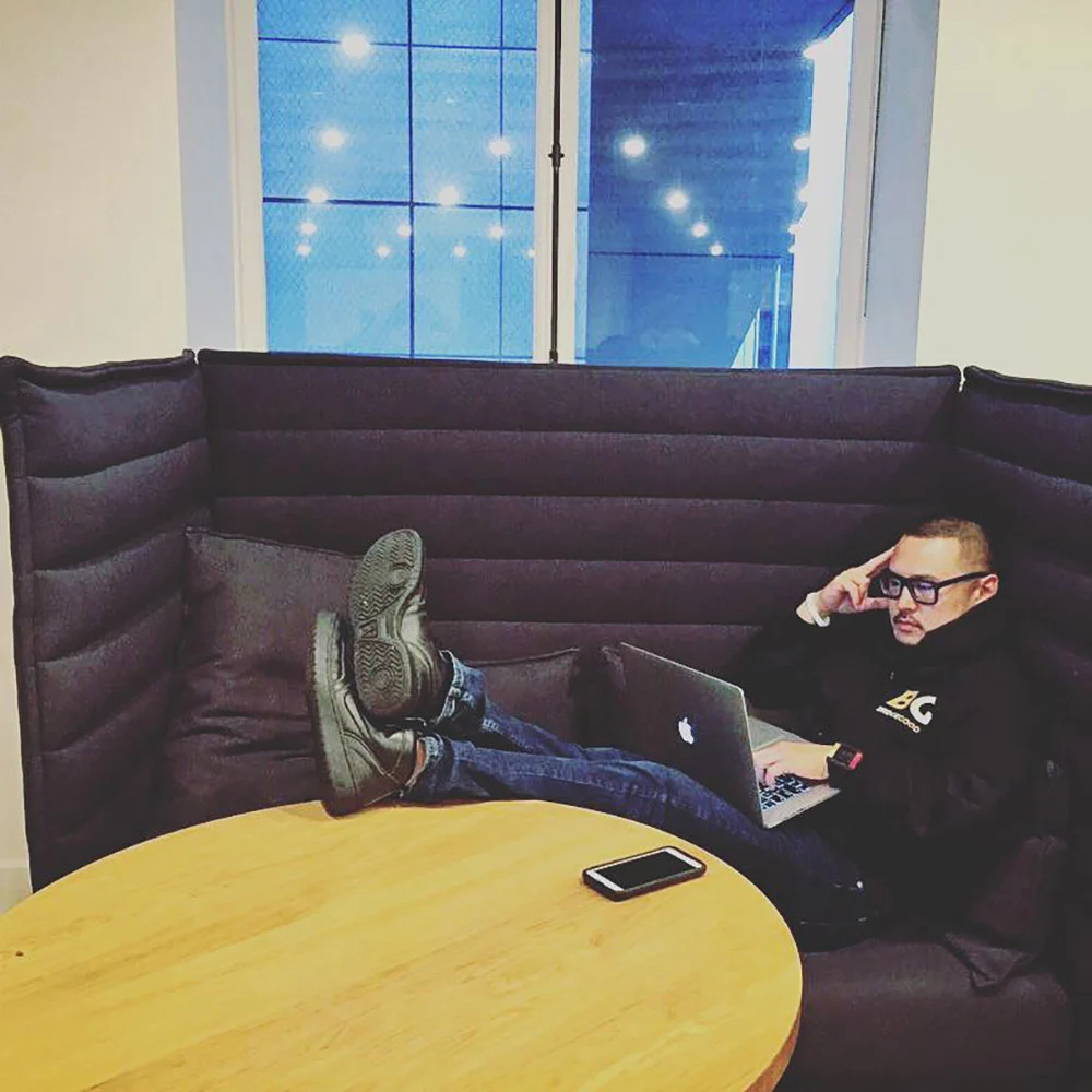 A man with glasses lounges in a padded booth, feet on the seat, using a laptop beside a wooden table.