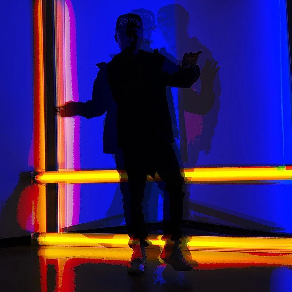 A person in dark clothing poses with arms out amid blue light, orange light bars and overlapping shadows.