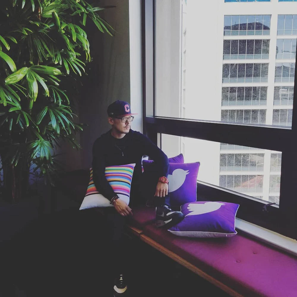 A person in a cap sits on a purple window bench with colorful pillows beside a tall plant and office windows.