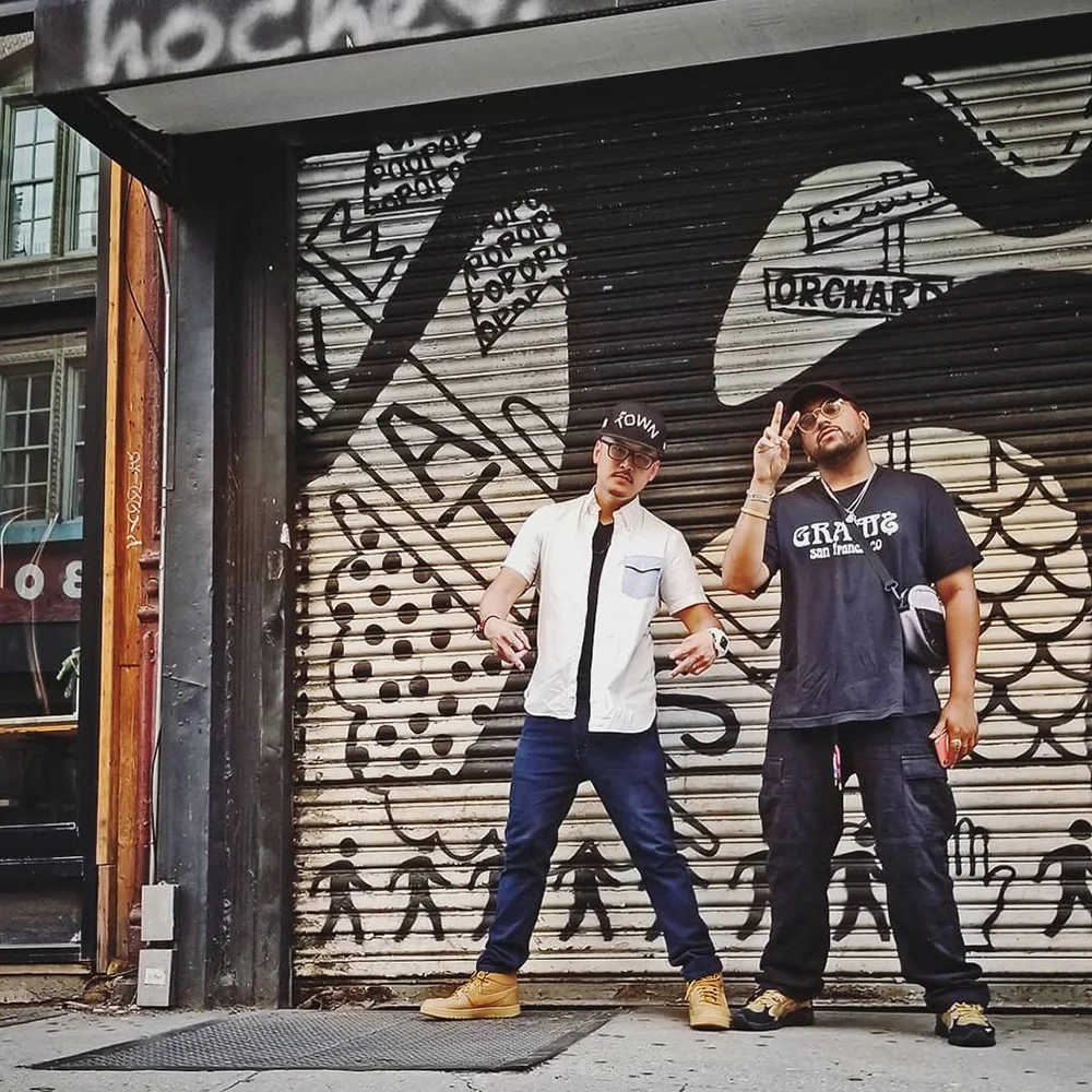 Two men pose on a sidewalk in front of a closed storefront shutter covered in black-and-white artwork.