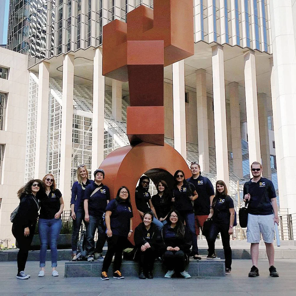 Twelve people pose around a large red geometric sculpture outside a modern building with tall white columns.