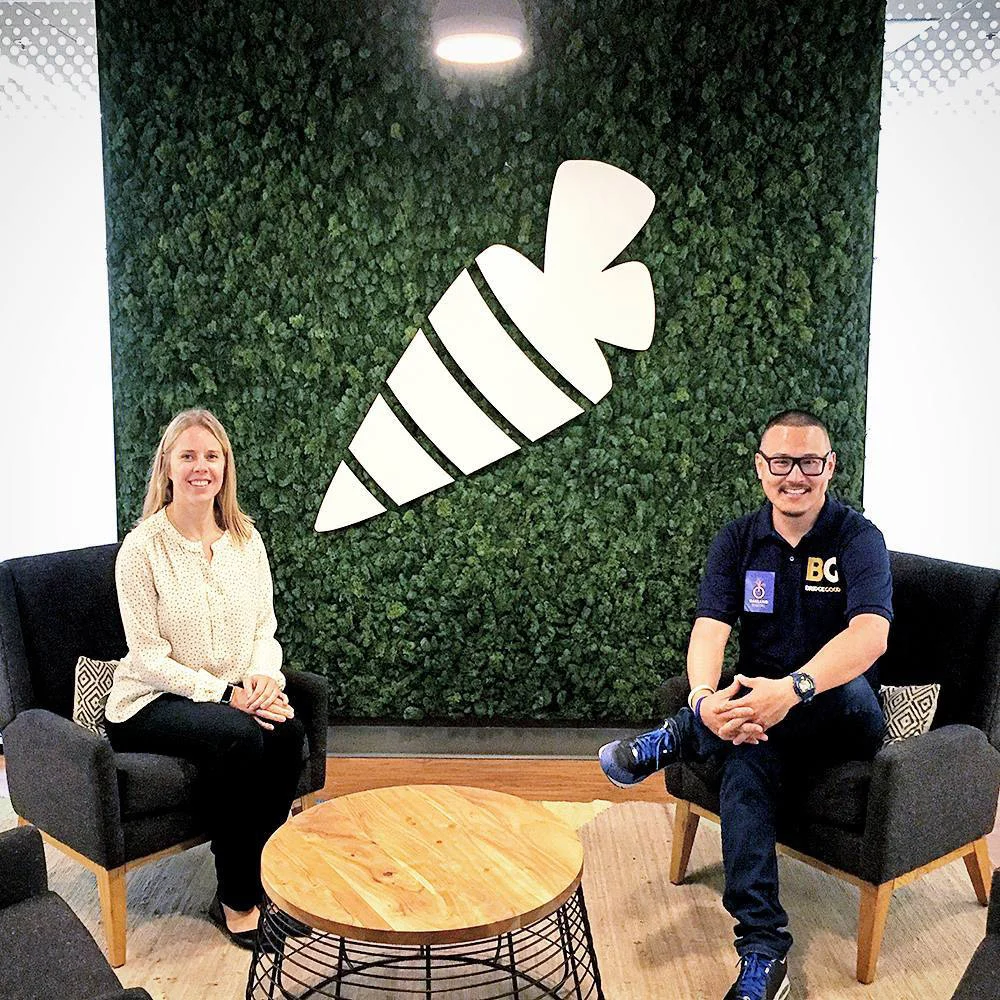 Two people sit in armchairs beside a round wooden table, with a large white carrot on a green plant wall behind them.