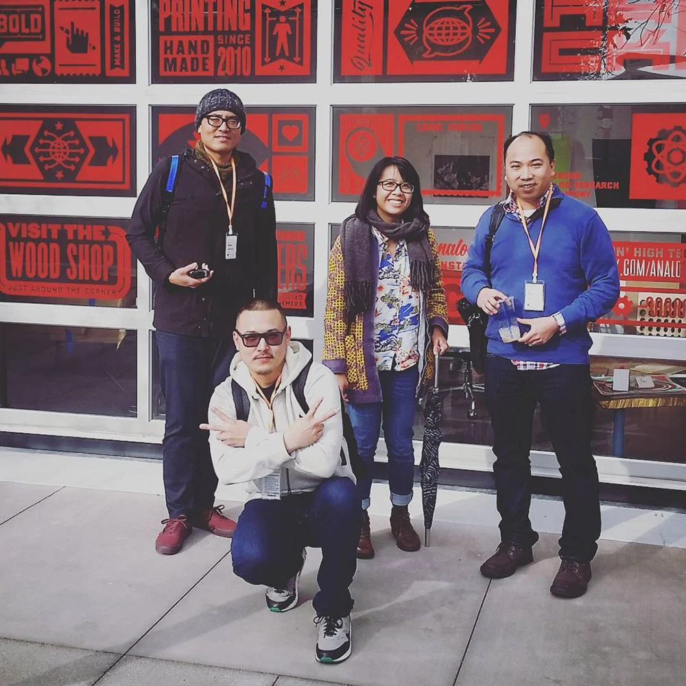 Four people wearing lanyards pose, one crouching, in front of windows filled with red and black printmaking posters.