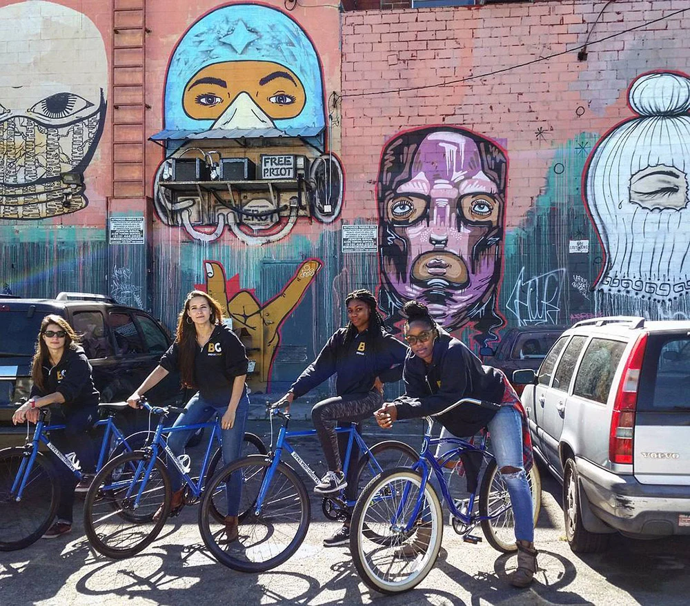 Four people pose with bicycles between parked cars in front of a brick wall covered in colorful portraits.
