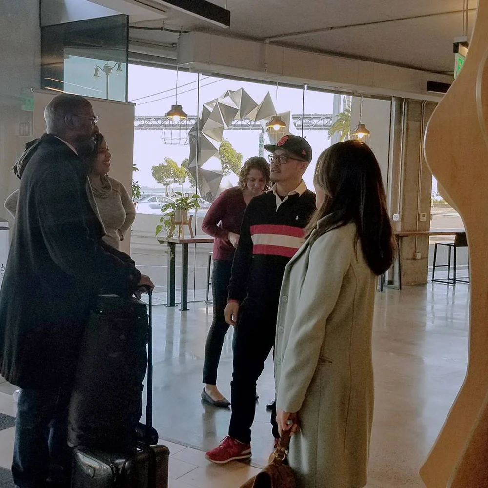 Five people gather and chat in a bright room with large windows, plants, and geometric hanging decor.