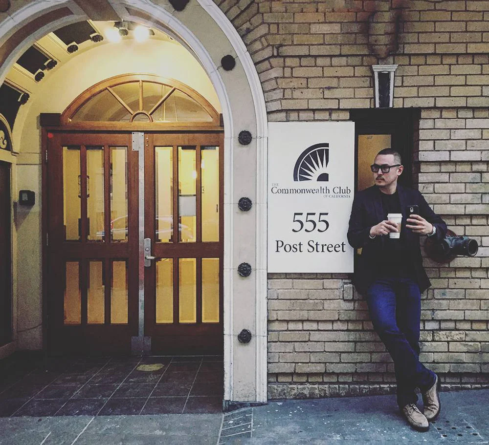 A man holding a cup leans against a brick wall beside the arched entrance to the Commonwealth Club at 555 Post Street.