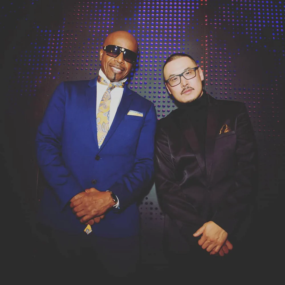 Two men in suits, one wearing sunglasses and one glasses, pose before a perforated wall lit blue and purple.