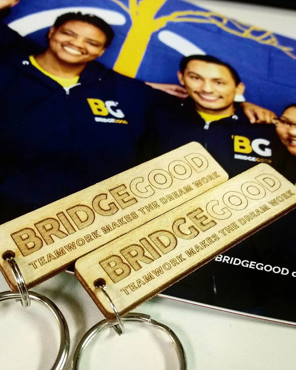 Wooden BRIDGEGOOD keychains rest on a printed photo of three smiling people with arms around each other.