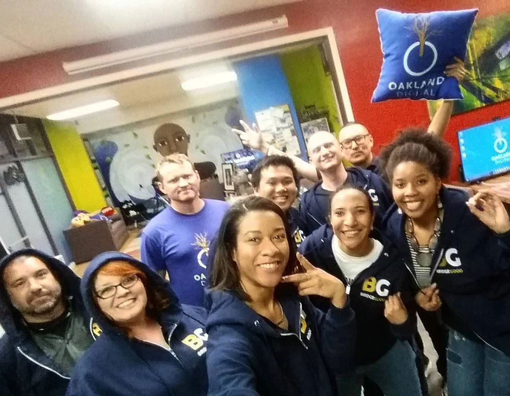 Nine people crowd together for a group selfie in an office, with one holding a blue Oakland Digital pillow overhead.