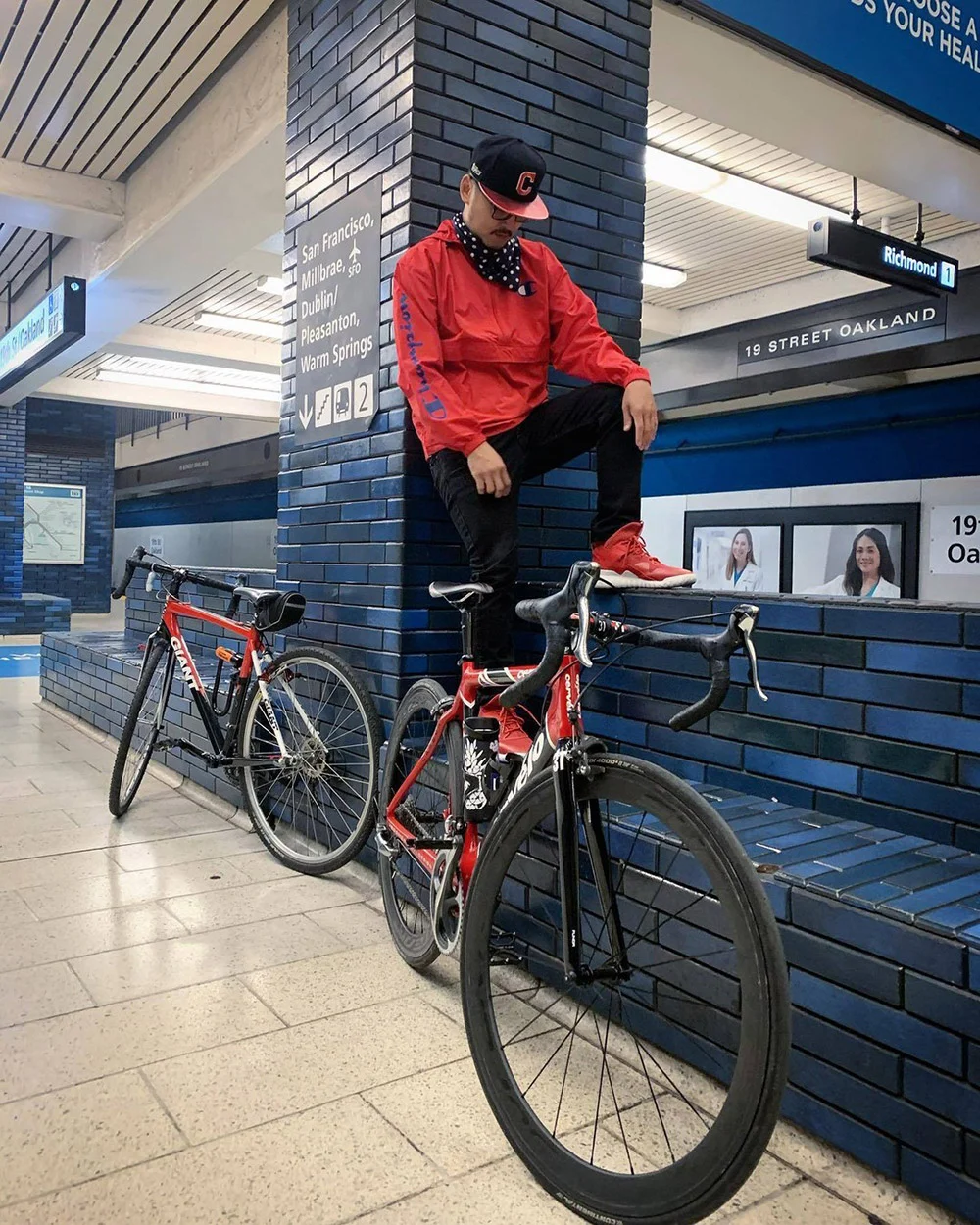 A person in a red jacket sits on a tiled ledge beside two bicycles at Oakland's 19th Street BART station.