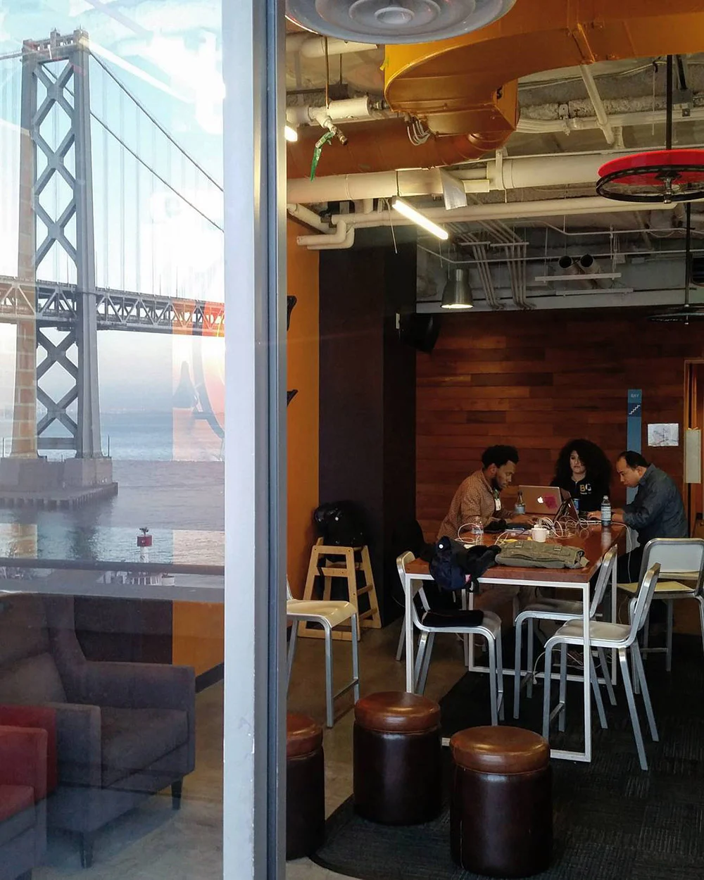 Three people sit around a table in a café with exposed ducts, beside a window reflecting the Bay Bridge.