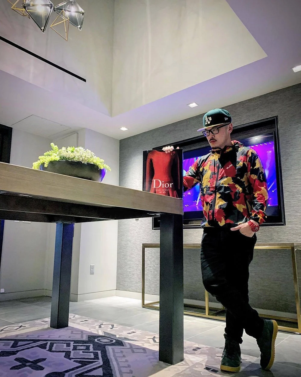 A person in a floral shirt and cap leans on a tall table beside a Dior bag in a modern room.