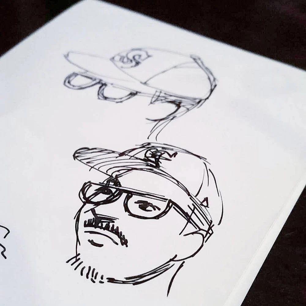 Two pen sketches on white paper show a person wearing glasses and a baseball cap, one with a mustache and beard.