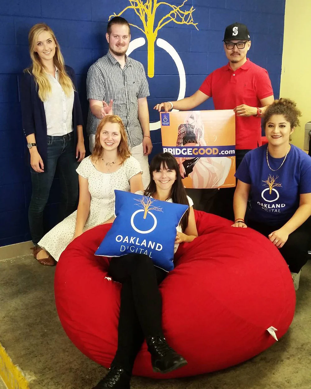Six people pose by a red beanbag against a blue wall, holding a BRIDGEGOOD sign and an Oakland Digital pillow.