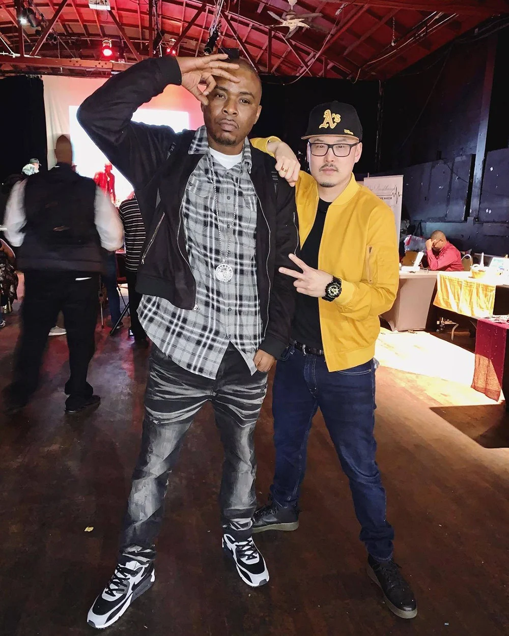 Two men pose with hand gestures in a hall with red ceiling beams, one wearing plaid and the other a yellow jacket.