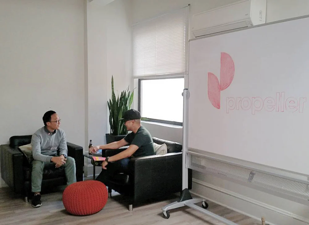 Two people sit facing each other in black armchairs beside a whiteboard with a pink Propeller logo.