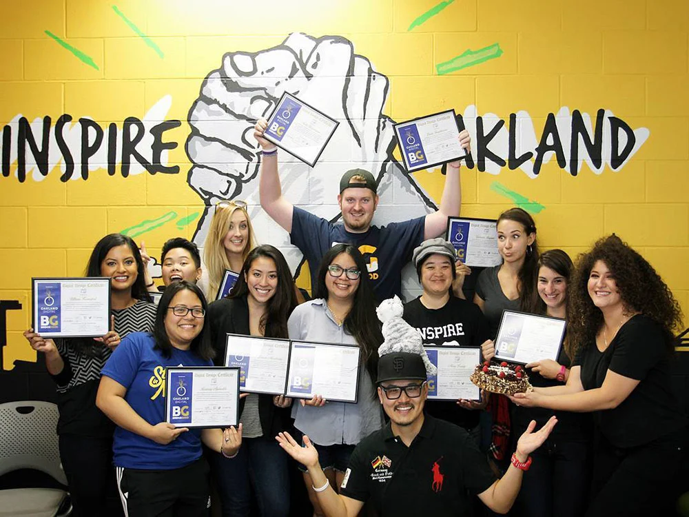 A group poses with certificates in front of a yellow wall with an Oakland mural, one person raising awards overhead.