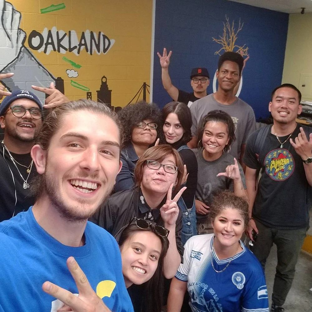 A group crowds together for a smiling selfie in front of a colorful Oakland mural, several flashing peace signs.