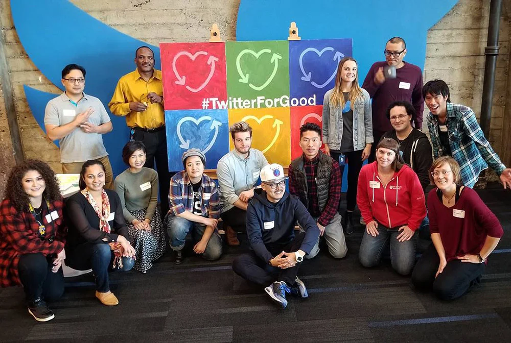 A group poses around a colorful #TwitterForGood display with heart symbols in a brick-walled room.