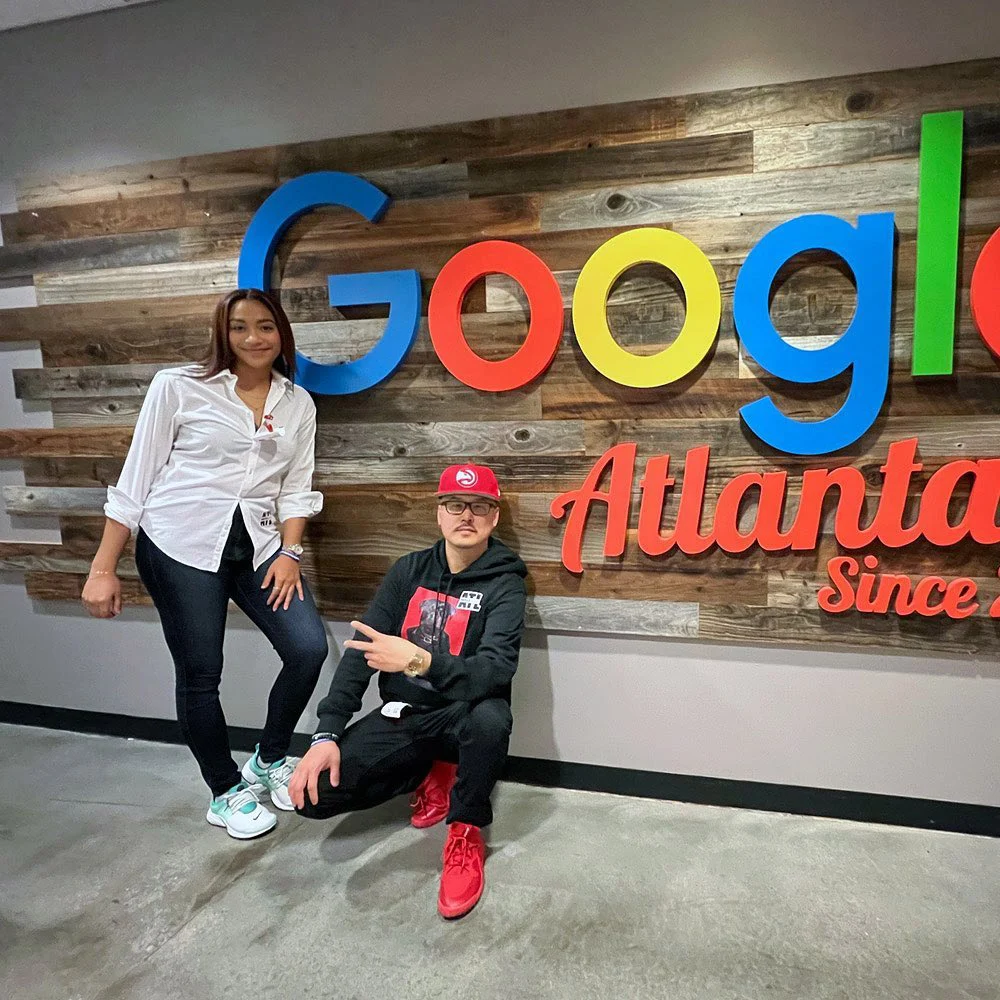 Two people pose by a Google Atlanta sign on a wood wall, one standing and one crouching with a peace sign.