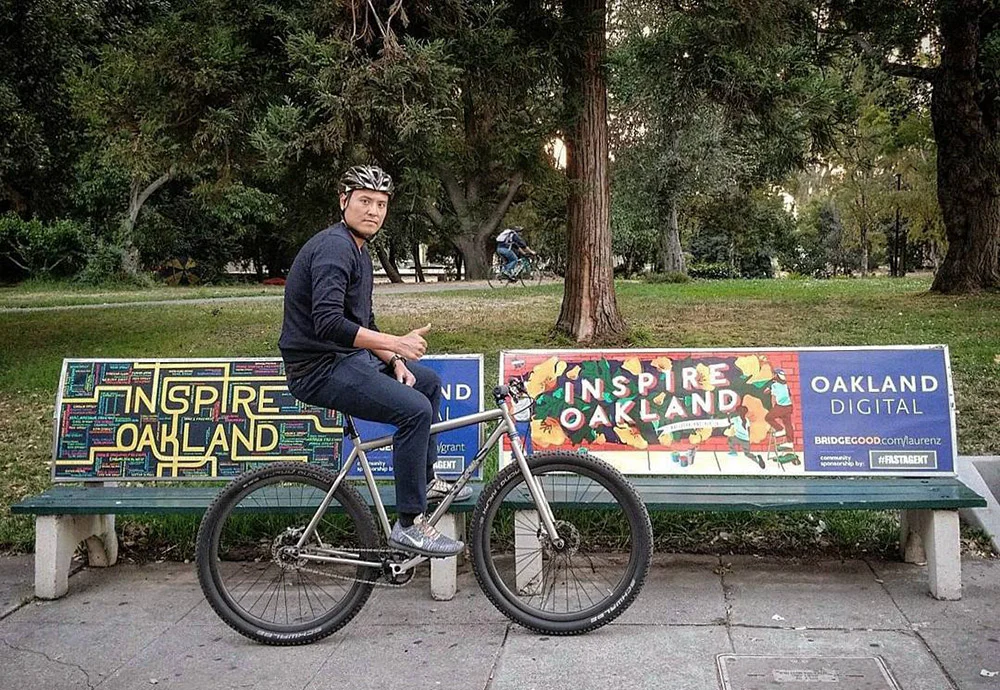 A helmeted cyclist sits on a bicycle in front of a colorful Inspire Oakland bench in a tree-filled park.