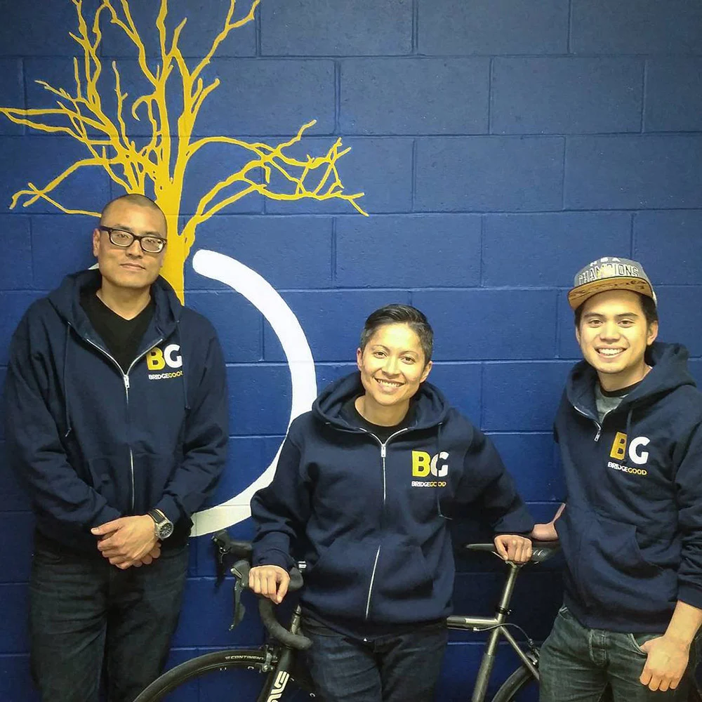 Three people in matching navy hoodies pose with a bicycle against a blue wall painted with a yellow tree.