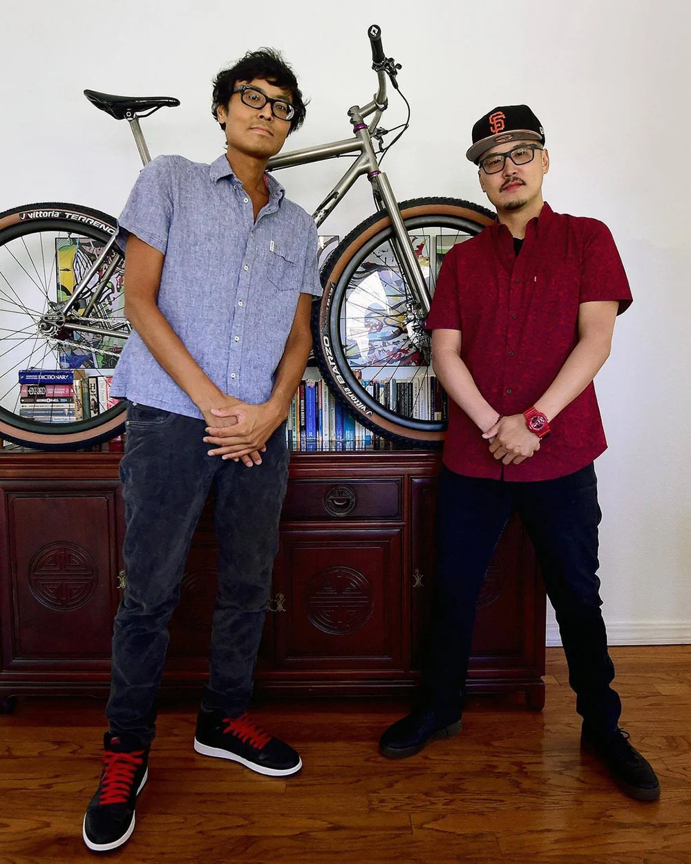 Two people stand with hands clasped in front of a bicycle displayed above a wooden cabinet lined with books.